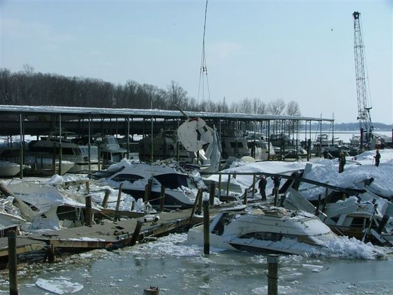 Boats damaged and covered in snow at a marina, with a crane in the background