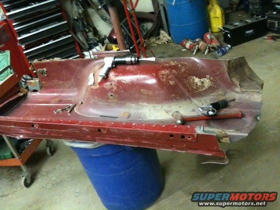 Red metal panel with extensive rust and damage, resting on a blue barrel. Tools are on the panel