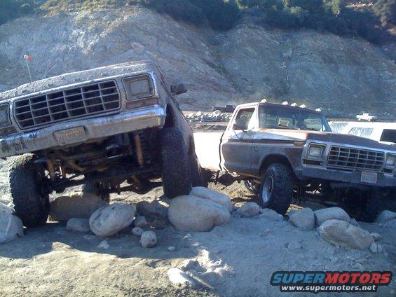 Two lifted Ford trucks navigating rocky terrain. The front truck is silver, the rear truck is brown