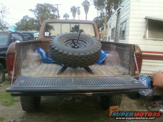 Brown pickup truck bed with a spare tire mounted. Other vehicles and a trailer are visible