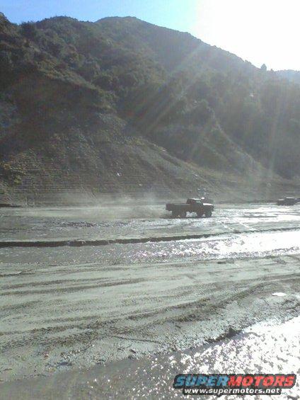 Side view of a 1976 Ford F-250 driving through a dusty, wet terrain with another truck in the background
