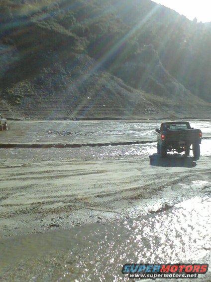 Pickup truck driving on a muddy, wet surface with a large hill in the background. Another vehicle is partially visible to the left