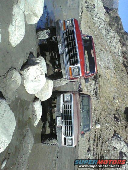 Two Ford F-Series trucks on a rocky incline; one red, one brown. The red truck is in front