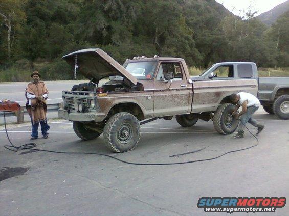 Brown Ford pickup with hood up, guy working on front tire. Another truck behind it