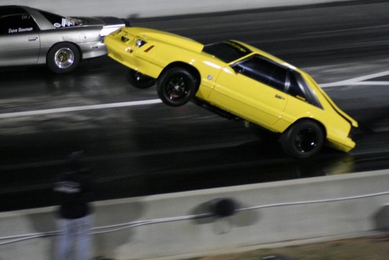Yellow fox body Mustang doing a wheelie on a drag strip next to a silver car