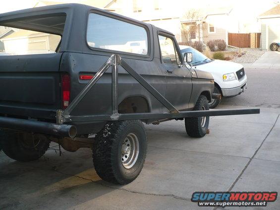 Rear view of a dark-colored Ford Bronco with a custom-welded tire carrier extension. A white Ford F-150 is parked behind it