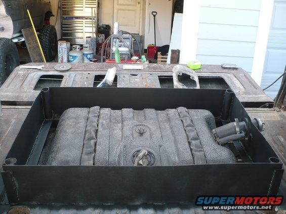 Aftermarket fuel tank mounted in a metal bracket inside a truck bed. A fuel pump is attached to the side of the tank