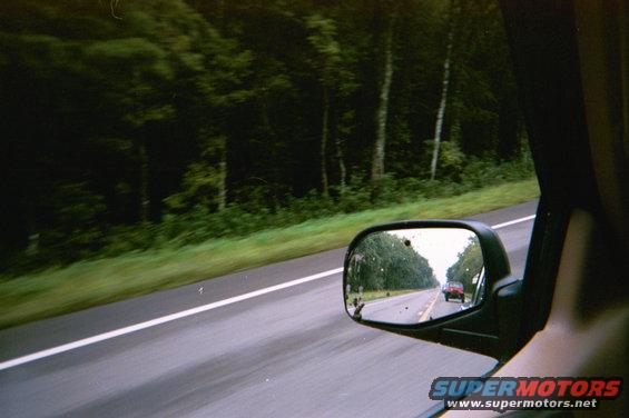 Red car seen in side mirror on a highway with trees lining the road