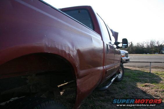 Red pickup truck with dents on the rear quarter panel, viewed from the rear quarter angle