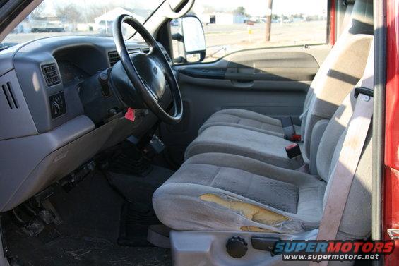 Truck interior with torn driver's seat, dashboard, steering wheel, and passenger seats. Red exterior visible