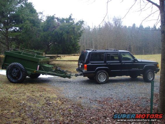 Black Jeep Cherokee with silver rims towing a green utility trailer on a gravel road