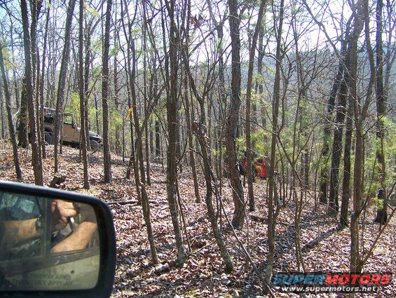 Tan Jeep Wrangler off-road in a wooded area, seen from side mirror