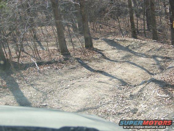 Dirt trail through woods with ruts and bumps. Part of a vehicle hood in foreground