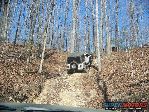 Jeep Wrangler driving up a wooded dirt trail, another vehicle visible in the distance