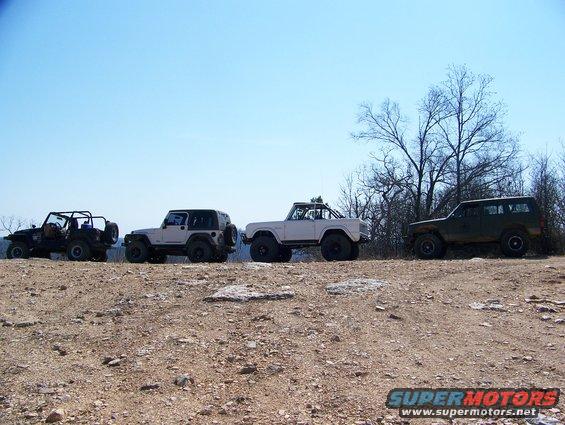 Four off-road vehicles, including Jeeps and a Bronco, parked on a dirt hill under a clear blue sky