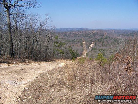 Dirt road winds through dry grass and trees, leading to a view of distant hills and power lines