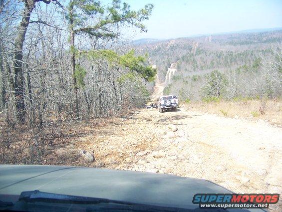 Two SUVs driving up a rocky, wooded trail. One is in front, the other is further back