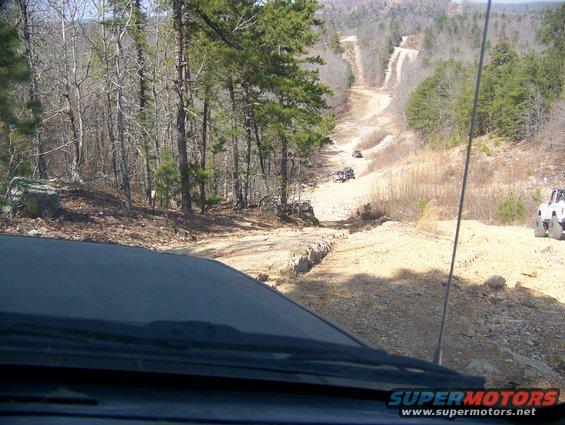 View from inside a vehicle looking down a rocky, wooded off-road trail with several other vehicles ahead and to the side