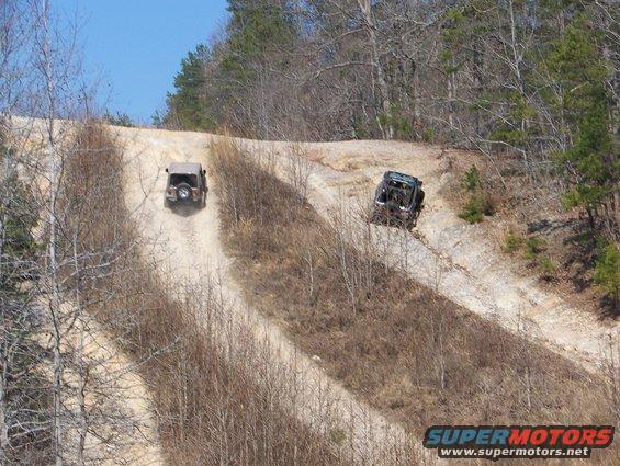 Two off-road vehicles ascend a steep dirt track in a wooded area