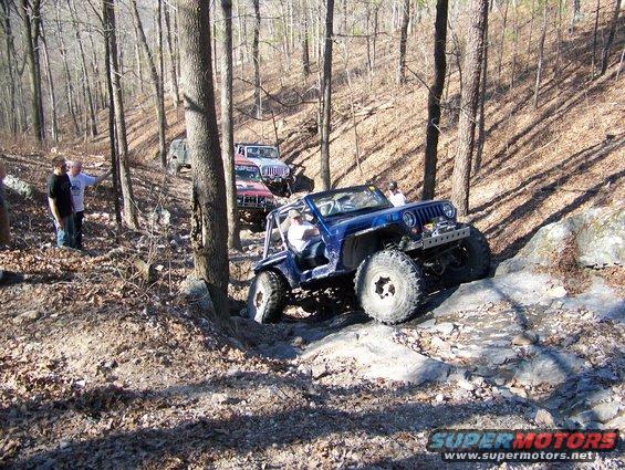 Several Jeeps navigating a rocky, wooded trail with people watching