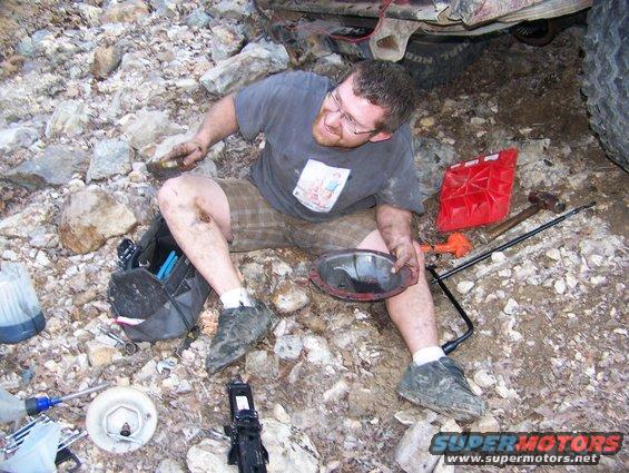 Man sitting on rocky ground, holding a differential cover, with tools and a red jerry can nearby. Part of a large tire and vehicle frame visible