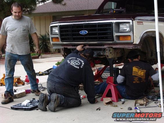 Maroon and tan Ford truck front end on jack stands, men working underneath. Visible Ford grille
