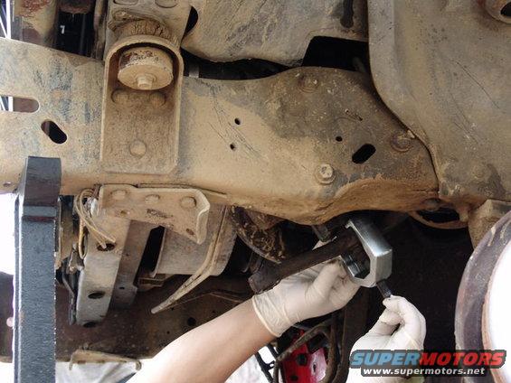Gloved hands use a tool on a vehicle's suspension components, likely during maintenance or repair