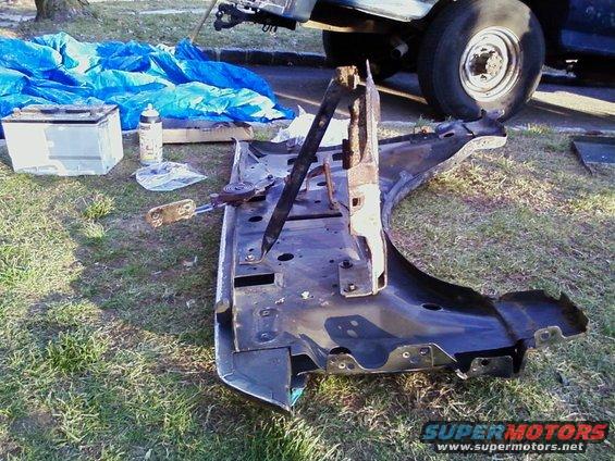 Car fender assembly with bracket on grass, blue tarp and battery nearby