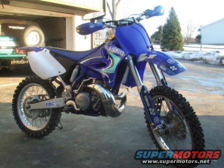 Blue Yamaha dirt bike parked in front of a garage. A green trailer is visible inside the garage