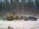2000 Chevrolet Tahoe - A dark pickup truck tows a flatbed trailer carrying a yellow tractor through a wooded landscape.
