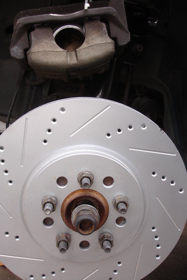 Brake caliper over a drilled and slotted rotor with five lug nuts visible