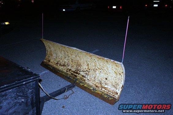 Rusty yellow snow plow with pink markers attached to a trailer hitch