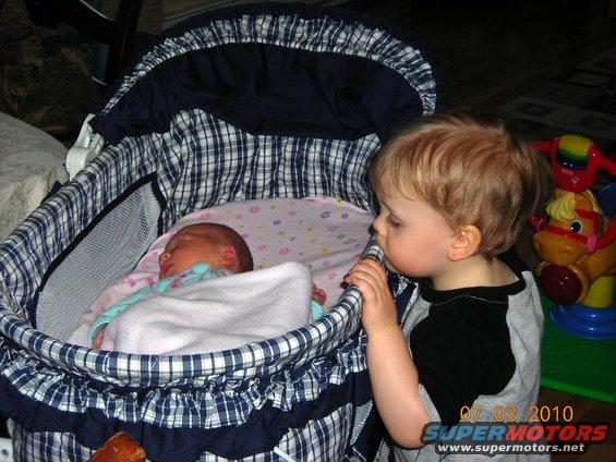 Toddler peers into a bassinet at a sleeping baby. Date stamp reads 07.03.2010