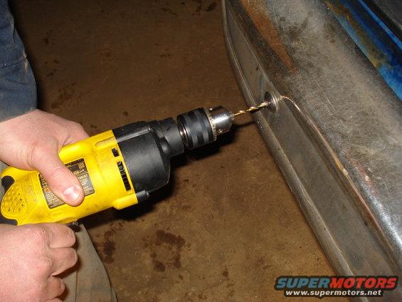 Yellow drill bit bores into a scratched chrome bumper, with blue paint visible behind