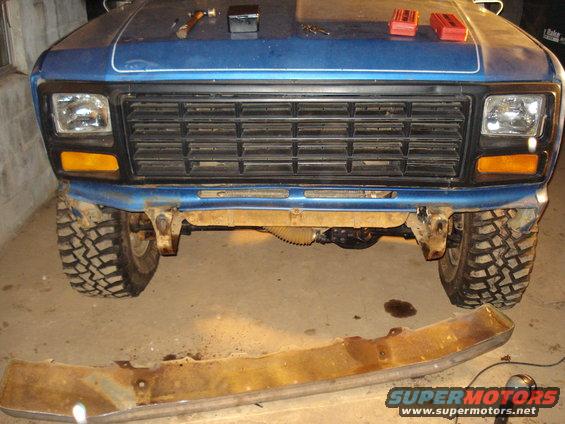 Blue pickup truck front end with a large grille and off-road tires. A rusty bumper lies on the ground