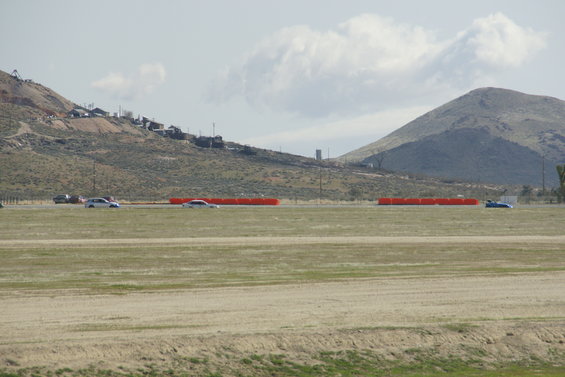 Several cars drive on a road with orange barriers, set against a backdrop of hills and buildings
