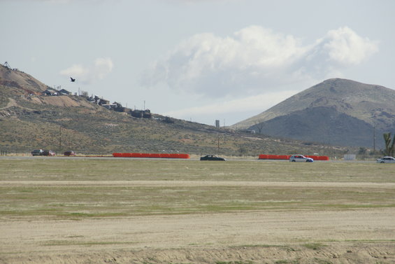 Cars driving on a track with orange barriers, hills, and buildings in background