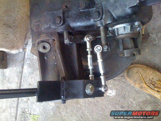 Transfer case linkage with adjustable rods and a black bracket. A tan boot is visible on the right
