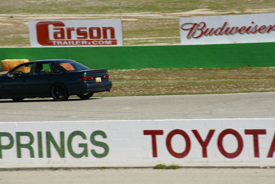 Dark sedan driving on a race track with sponsor signs in the background