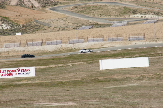 White sedan and black sedan on a race track, with grandstands and a banner in the foreground