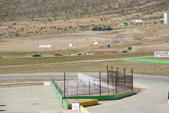 Several cars drive on a racetrack with grandstands and dry hills in the background