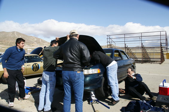 Several people are gathered around the open hood of a dark sedan, appearing to work on it. Another car with the number 07 is parked nearby