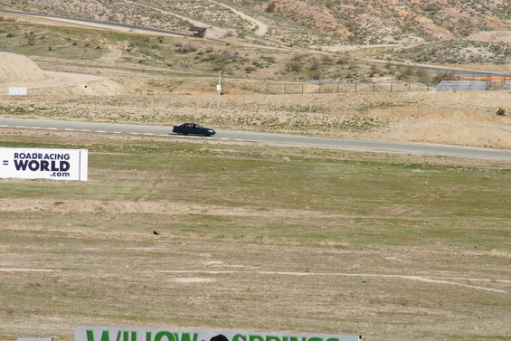 Black sedan on a race track, side view. Roadracingworld.com sign visible