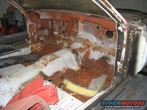 1970 Ford Mustang interior, heavily rusted, showing floor pan and firewall, with red plastic parts on the floor
