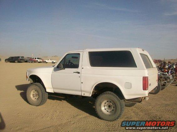 White SUV parked on sand, side view. Visible are large tires, tinted windows, and a spare tire mount on the rear