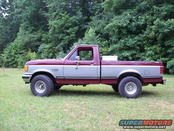 Side view of a maroon and silver Ford F-150 pickup truck parked on grass with trees in the background. It has "F-150" badging on the fender