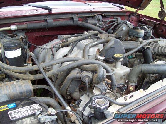 Engine bay with hoses, wires, and engine components visible. Battery in foreground