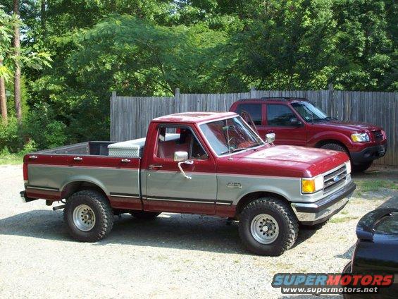 1990 Ford F-150 pickup truck, two-tone red and silver, with a toolbox in the bed. Red SUV parked behind it