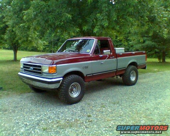 Two-tone red and gray Ford F-150 pickup truck with aftermarket wheels and tires, parked on a gravel driveway