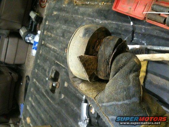 Gloved hand holding a rusted, cut section of exhaust pipe. Toolbox and other tools visible in background